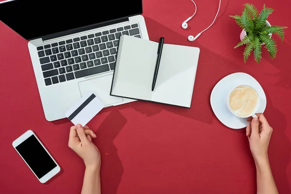 Vista parcial de la mujer que sostiene la tarjeta de crédito y la taza de café cerca de teléfono inteligente, ordenador portátil, auriculares, portátil y planta sobre fondo rojo - foto de stock