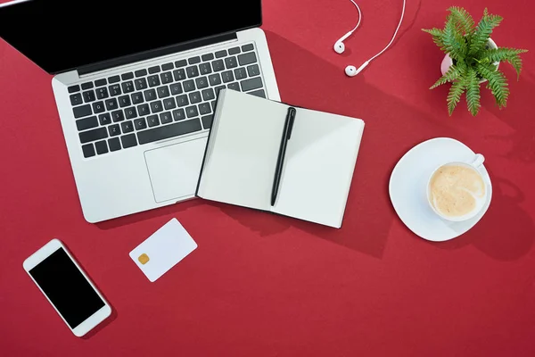 Top view of credit card on red background with smartphone, laptop, earphones, coffee, notebook and plant — Stock Photo