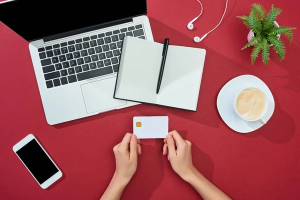 Vista parcial de la mujer que sostiene la tarjeta de crédito cerca de la taza de café, teléfono inteligente, computadora portátil, auriculares, portátil y planta sobre fondo rojo - foto de stock