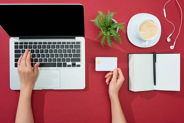 Обрезанный вид женщины, держащей кредитную карту и с помощью ноутбука рядом наушники, кофе, ноутбук с ручкой и завод на красном фоне — стоковое фото