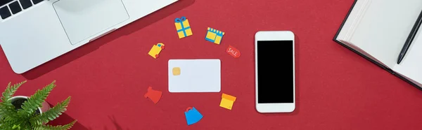 Vista superior de la tarjeta de crédito con iconos sobre fondo rojo con teléfono inteligente, ordenador portátil, portátil y planta, plano panorámico - foto de stock