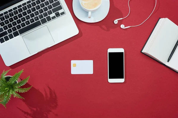 Vista dall'alto di carta di credito, smartphone, laptop, auricolari, caffè, notebook e piante su sfondo rosso — Foto stock