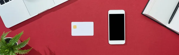 Vista superior de la tarjeta de crédito, teléfono inteligente, ordenador portátil, portátil y planta sobre fondo rojo, plano panorámico - foto de stock
