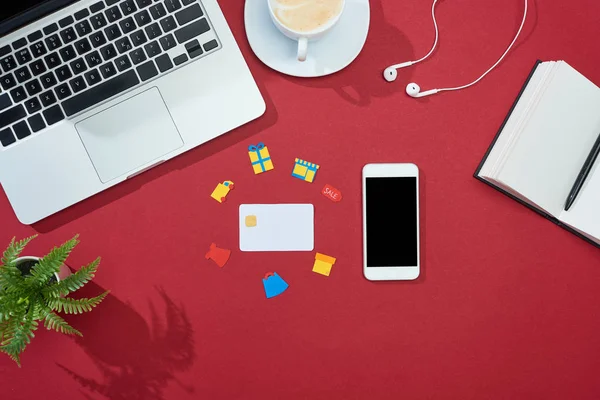 Top view of credit card with icons on red background with smartphone, laptop, earphones, coffee, notebook and plant — Stock Photo