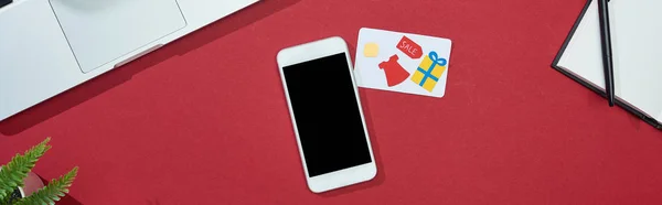 Vista superior de la tarjeta de crédito sobre fondo rojo con teléfono inteligente, ordenador portátil, bloc de notas, plano panorámico - foto de stock