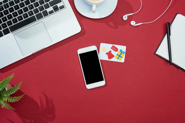 Top view of credit card on red background with smartphone, laptop, earphones and coffee — Stock Photo