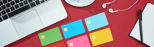Вид зверху на різнокольорові порожні кредитні картки на червоному тлі з ноутбуком, навушниками та блокнотом, панорамний знімок — стокове фото