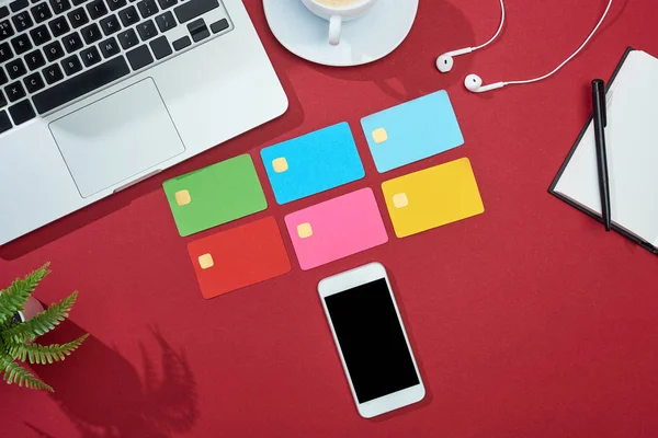 Vista superior de tarjetas de crédito vacías multicolores sobre fondo rojo con teléfono inteligente, portátil, auriculares y café - foto de stock
