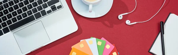 Vista superior de tarjetas de crédito vacías multicolores sobre fondo rojo con ordenador portátil, auriculares y café, plano panorámico - foto de stock
