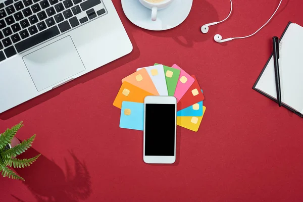 Вид сверху на многоцветные пустые кредитные карты на красном фоне со смартфоном, ноутбуком, наушниками и кофе — Stock Photo