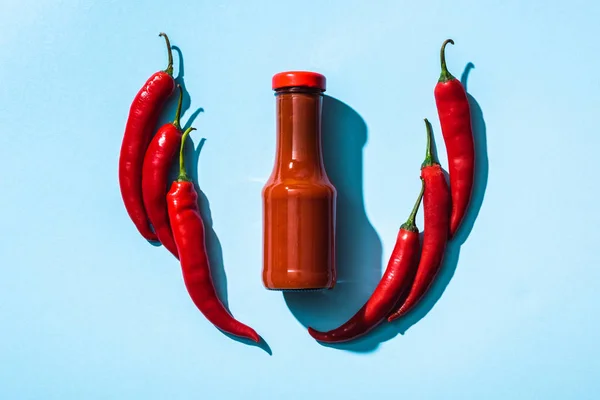 Vue du dessus de la sauce chili en bouteille entre les piments sur fond bleu — Photo de stock