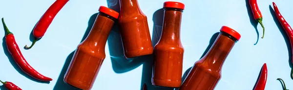 Top view of bottles with chili sauce beside chili peppers on blue surface, panoramic shot — Stock Photo