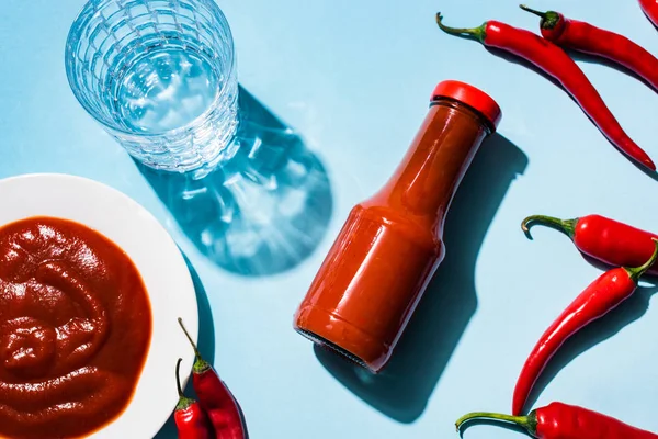 Vue du dessus de la sauce chili en bouteille et assiette avec piments et verre d'eau sur fond bleu — Photo de stock