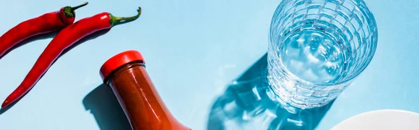 Top view of glass of water with chili sauce and chili peppers on blue background, panoramic shot — Stock Photo