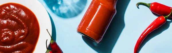 Top view of chili peppers with tasty ketchup in pale and bottle on blue surface, panoramic shot — Stock Photo