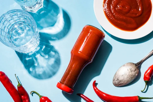 Top view of glasses of water with chili sauce in plate and bottle beside chili peppers on blue background — Stock Photo