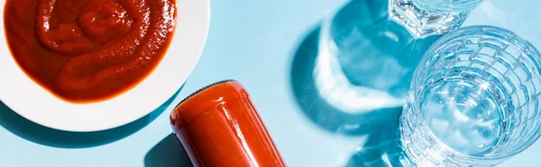 Top view of homemade tomato sauce in plate and bottle beside glasses of water on blue background, panoramic shot — Stock Photo