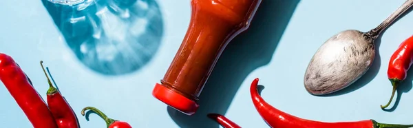 Top view of bottle with chili sauce, spoon and chili peppers on blue surface, panoramic shot — Stock Photo