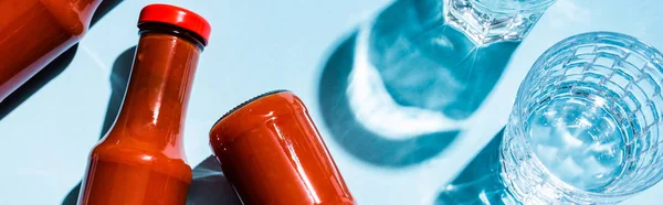 Vista dall'alto di bottiglie con gustoso ketchup accanto a bicchieri d'acqua su sfondo blu, colpo panoramico — Foto stock