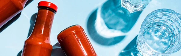 Vista superior de botellas con sabroso ketchup junto a vasos de agua sobre fondo azul, plano panorámico — Stock Photo
