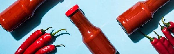 Vista superior de molho de pimenta caseiro em garrafas com pimentas na superfície azul, tiro panorâmico — Fotografia de Stock
