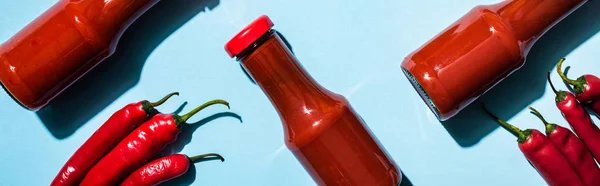 Top view of homemade chili sauce in bottles with chili peppers on blue surface, panoramic shot — Stock Photo