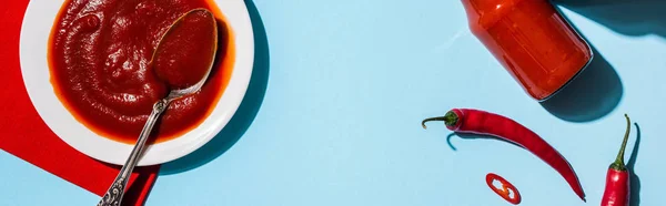 Top view of spicy tomato sauce in plate and bottle with chili peppers on blue surface, panoramic shot — Stock Photo