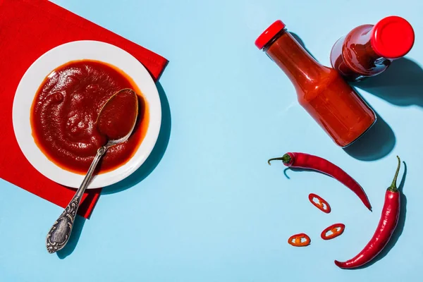 Ansicht der hausgemachten Chilisoße in Flaschen und Teller mit reifen Chilischoten auf blauer Oberfläche — Stockfoto