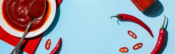 Vista superior do saboroso molho de pimenta na placa ao lado de pimentas com fatias no fundo azul, tiro panorâmico — Fotografia de Stock