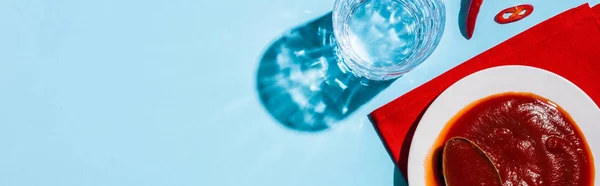 Top view of chili sauce on plate beside glass of water and chili pepper on blue surface, panoramic shot — Stock Photo