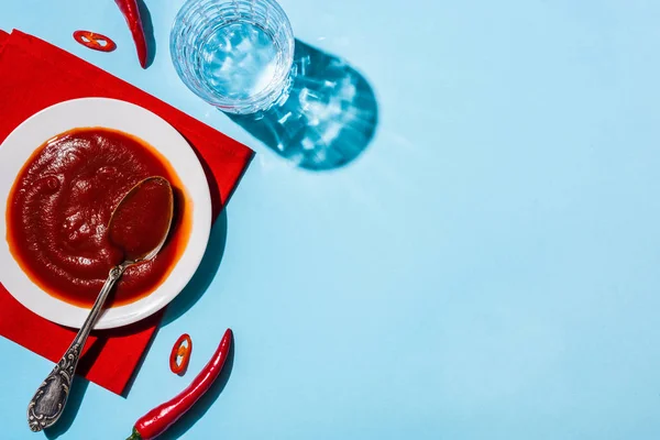 Köstliche Chilisoße in Pastete auf roter Serviette neben Glas Wasser und Chilischoten auf blauer Oberfläche — Stockfoto