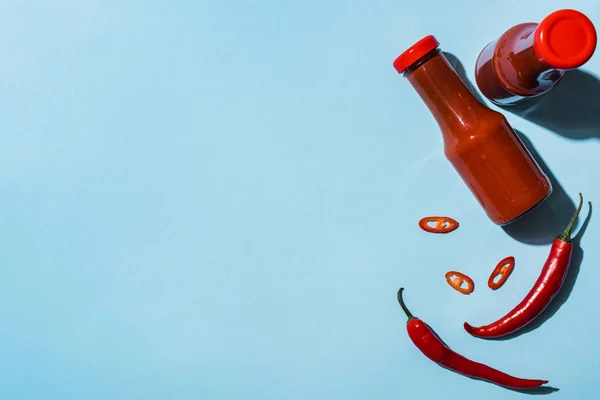 Vista superior de botellas con salsa de tomate picante junto a chiles sobre fondo azul - foto de stock