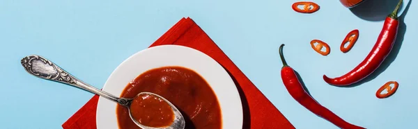 Top view of delicious spicy ketchup in plate with ripe chili peppers on blue surface, panoramic shot — Stock Photo