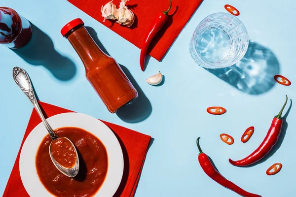 Top view of tasty tomato sauce with garlic and chili pepper beside glass of water on blue surface — Stock Photo