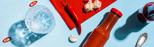 Top view of bottles with ketchup, garlic with chili pepper and glass of water on blue sutface, panoramic shot — Stock Photo