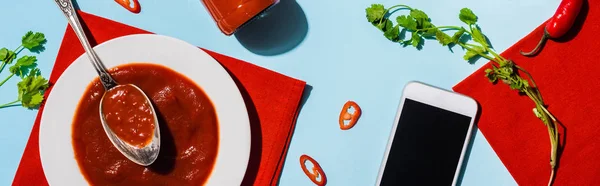 Top view of ketchup with chili pepper and cilantro beside smartphone with blank screen on blue surface, panoramic shot — Stock Photo