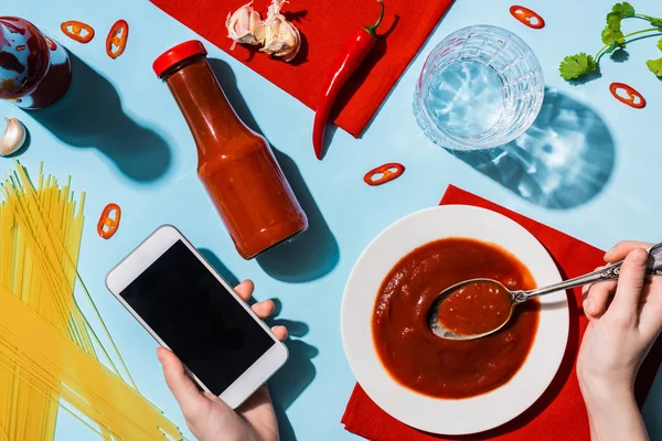 Vista recortada de la mujer sosteniendo el teléfono inteligente junto a ketchup, espaguetis y vaso de agua en la superficie azul - foto de stock