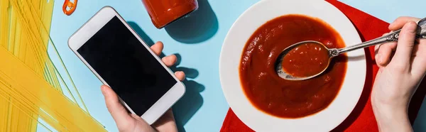 Cropped view of woman holding smartphone beside ketchup and spaghetti on blue background, panoramic shot — Stock Photo