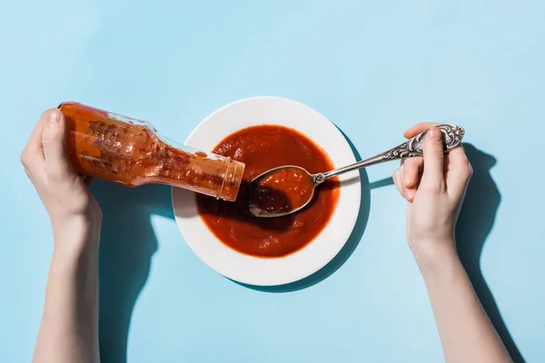 Vista recortada de la mujer sosteniendo cuchara y haciendo pucheros deliciosa salsa de tomate en plato sobre fondo azul - foto de stock