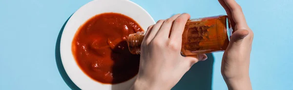 Vista cortada da mulher derramando ketchup saboroso de garrafa para placa na superfície azul, tiro panorâmico — Fotografia de Stock