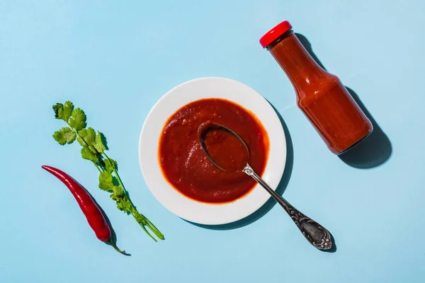 Vista dall'alto di salsa di pomodoro piccante in piatto e bottiglia con peperoncino e coriandolo su sfondo blu — Foto stock