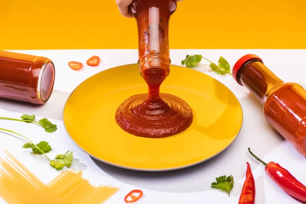 Vista recortada de la mujer vertiendo ketchup en el plato junto a espaguetis y chiles en la superficie blanca aislada en amarillo - foto de stock