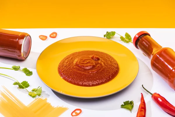 Homemade tomato sauce on plate beside spaghetti and chili peppers on white surface on yellow background — Stock Photo