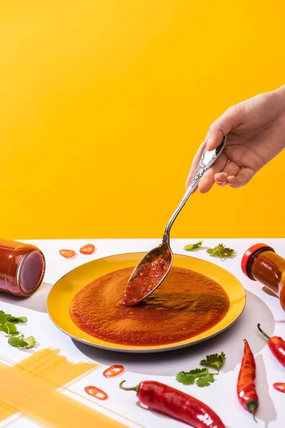 Cropped view of woman holding spoon with ketchup with spaghetti and chili peppers on white surface isolated on yellow — Stock Photo