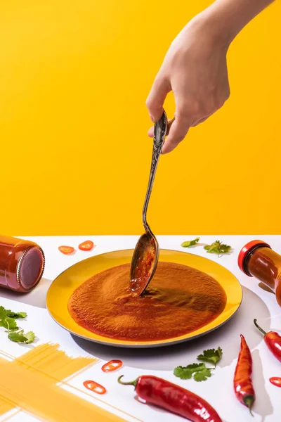 Vista recortada de la mujer sosteniendo cuchara por encima de la placa con salsa de tomate al lado de espaguetis en la superficie blanca aislada en amarillo - foto de stock