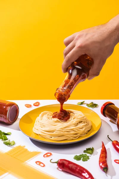 Обрізаний вид людини, що поливає кетчуп на спагеті з перець чилі та зеленню на білій поверхні ізольовані на жовтій — стокове фото