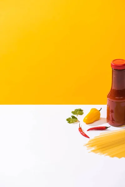 Garrafa de ketchup saboroso com pimentas e espaguete cru na superfície branca no fundo amarelo — Fotografia de Stock
