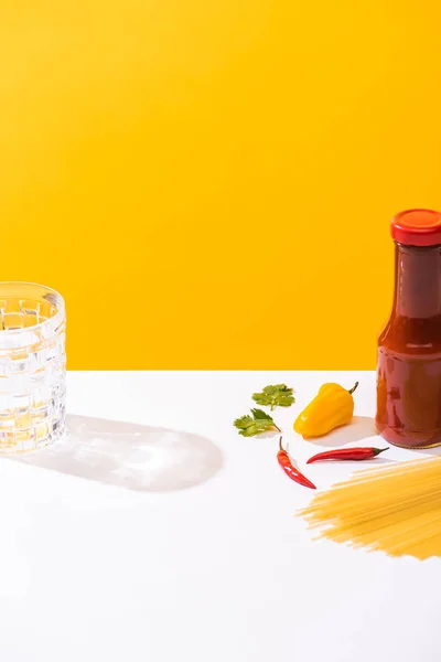 Bottle of ketchup beside peppers, raw spaghetti and glass of water on white surface on yellow background — Stock Photo
