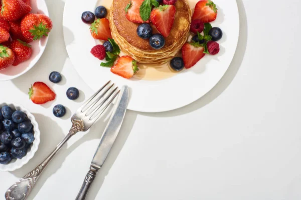 Top view of delicious pancakes with honey, blueberries and strawberries on plate with cutlery on marble white surface — Stock Photo