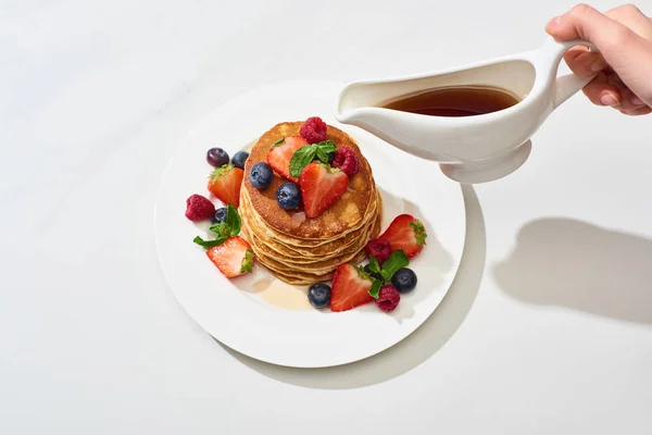 Cropped view of woman pouring maple syrup on delicious pancakes with blueberries and strawberries on plate on marble white surface — Stock Photo